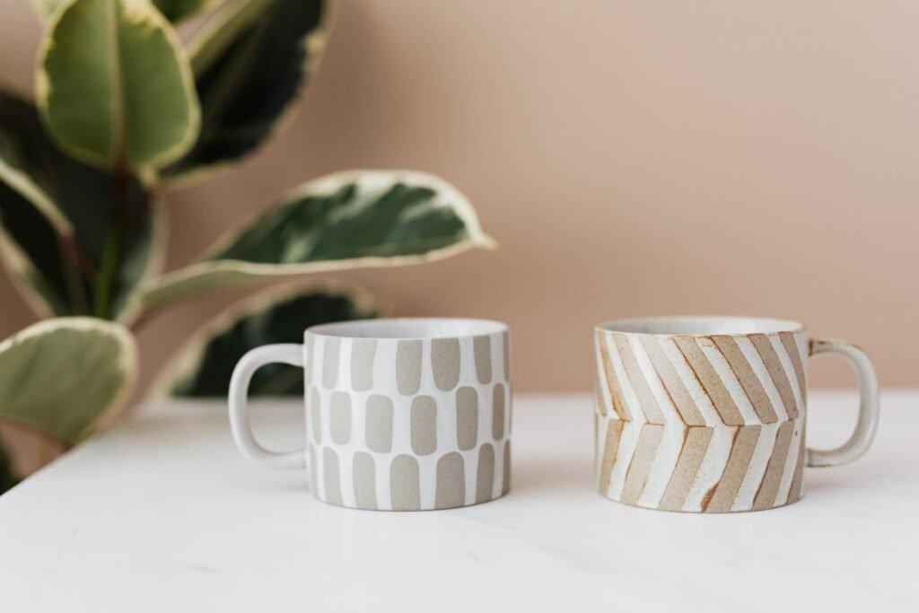 Handmade ceramic mugs with creative designs placed on white marble table with blurred green house plant near pink wall in background